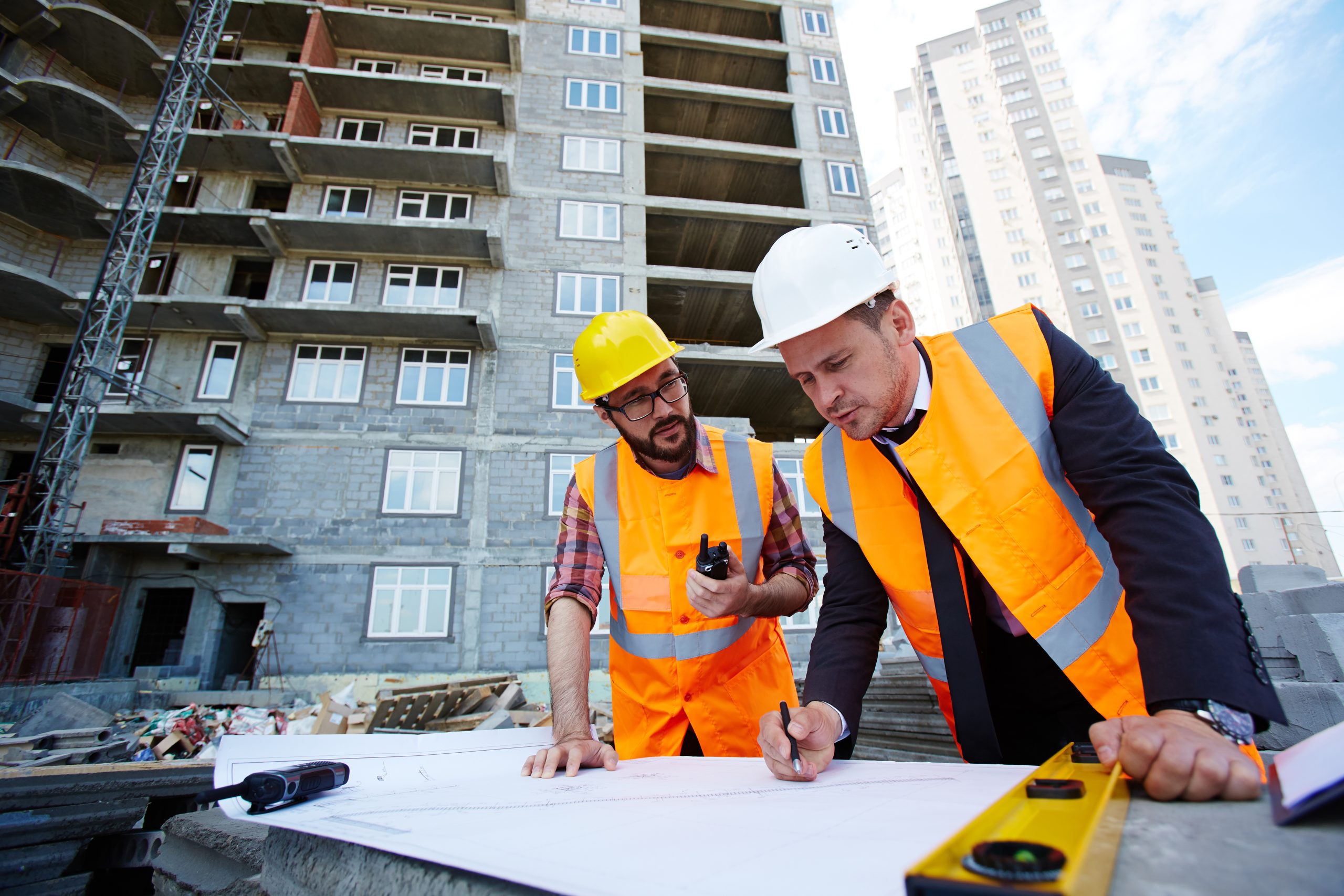 Pointing at sketch 2 construction contractors looking at plans with a tall building behind them
